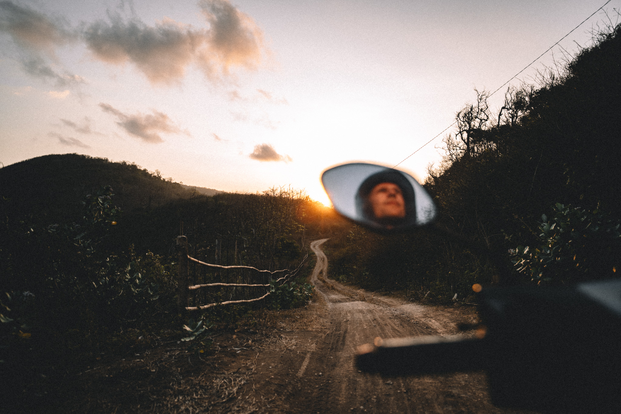 A face reflected in a scooter mirror beside a winding dirt road at sunset in Sumbawa